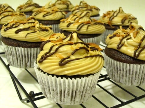 Samoas-inspired chocolate cupcakes topped with caramel frosting, toasted coconut, and chocolate drizzle, cooling on a wire rack.