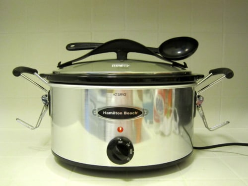 Hamilton Beach slow cooker with lid and serving spoon on top, set up on a kitchen counter.