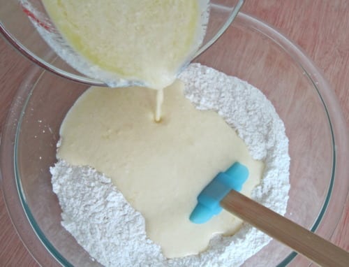 Wet ingredients being poured into a bowl of dry ingredients with a blue spatula, prepared for muffin batter during a blog anniversary recipe.