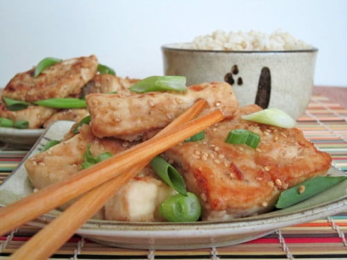 Sesame-crusted tofu topped with chopped green onions, served on a plate with chopsticks and a side of brown rice in the background.
