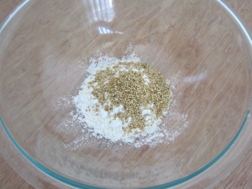 Flour and sesame seeds combined in a clear glass bowl on a wooden surface, prepared for coating tofu.