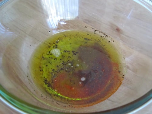 Olive oil mixed with spices and seasoning in a clear glass bowl on a wooden surface, prepared for a quinoa salad dressing.