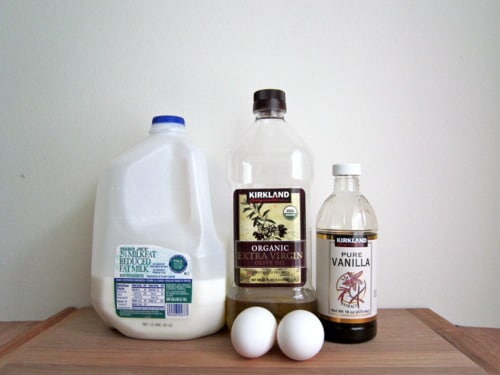 Milk, extra virgin olive oil, pure vanilla extract, and two eggs on a wooden surface.