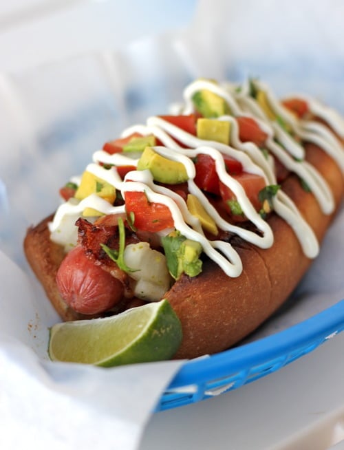Bacon-wrapped Sonoran hot dog topped with avocado, tomato, onion, and drizzled with crema, served with a lime wedge.