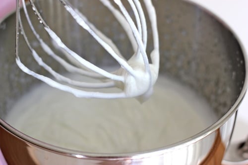Freshly whipped cream with soft peaks on a whisk attachment above a mixing bowl, prepared for strawberry shortcake muffins.
