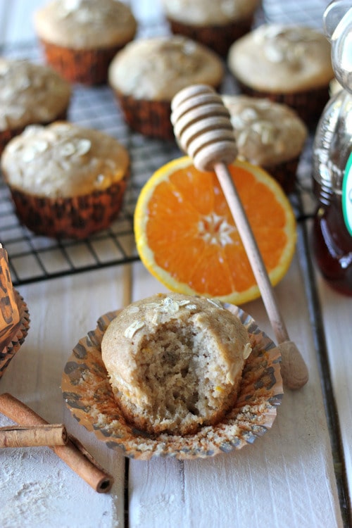 Orange cinnamon honey muffin with a bite taken out, surrounded by whole muffins, a halved orange, honey dipper, cinnamon sticks, and a cooling rack in the background.