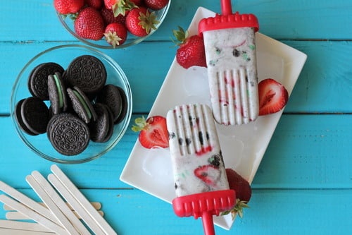 Strawberry mint cookies and cream popsicles on a white plate, surrounded by fresh strawberries and chocolate sandwich cookies.