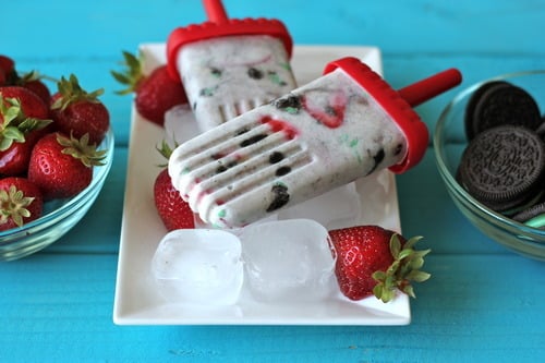 Strawberry mint cookies and cream popsicles on a plate with ice cubes, surrounded by fresh strawberries and chocolate sandwich cookies.