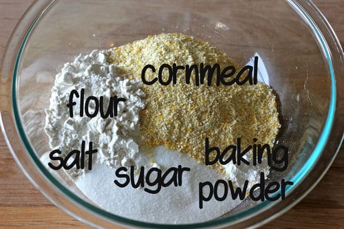 Glass bowl with labeled dry ingredients for blueberry cornbread muffins, including flour, cornmeal, baking powder, sugar, and salt.