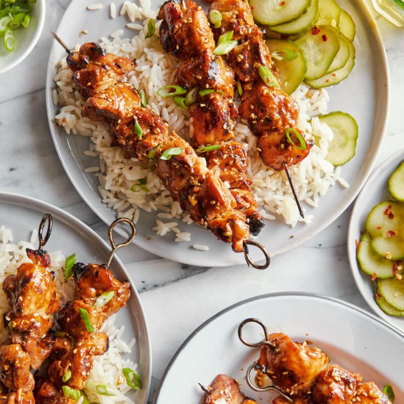 Plates of marinaded, sesame chicken kabobs, served on a bed of white rice with marinated cucumbers.