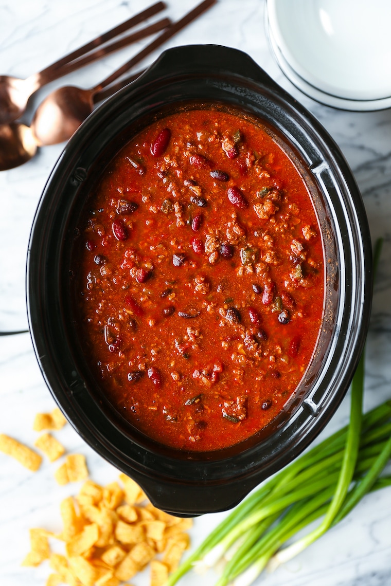 Slow cooker filled with hearty chili made with ground beef, kidney beans, black beans, and tomatoes, surrounded by green onions and corn chips.